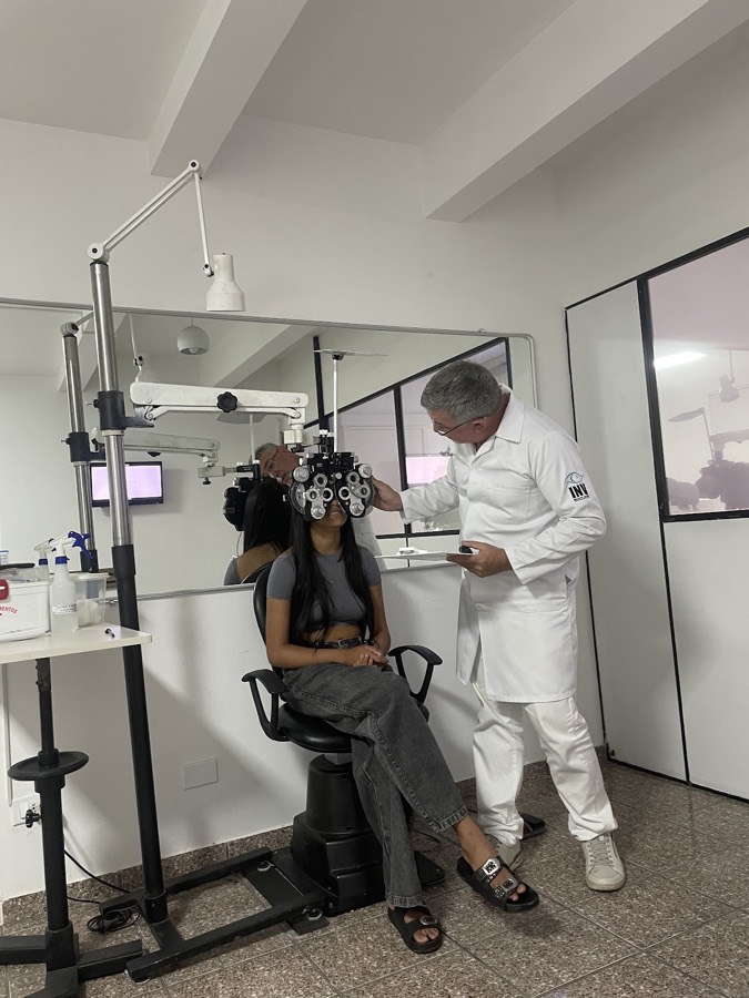 Celso Nunes, de jaleco branco, realizando exame de vista no foróptero com uma paciente
